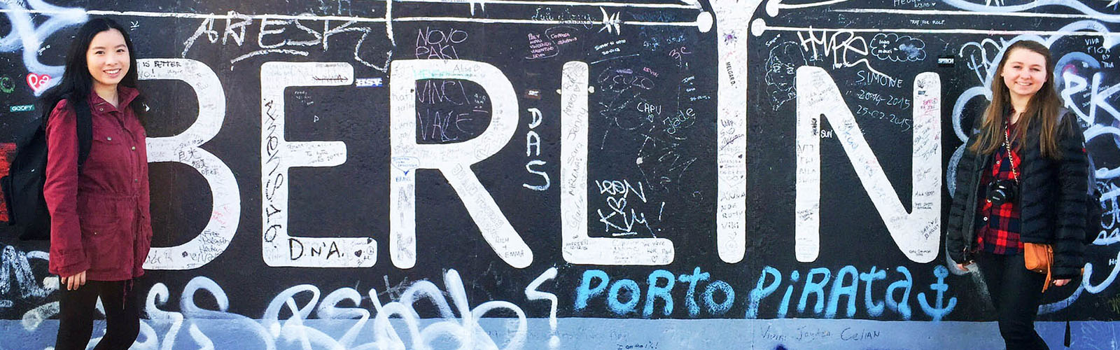 Two students are posed in front of a section of the Berlin wall. They are surround by graffiti and smiling at the camera.